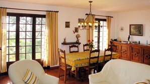 Zona pranzo con tavolo in legno, sedie e lampadario. Sono presenti finestre con tende, un armadietto in legno e un quadro appeso al muro.