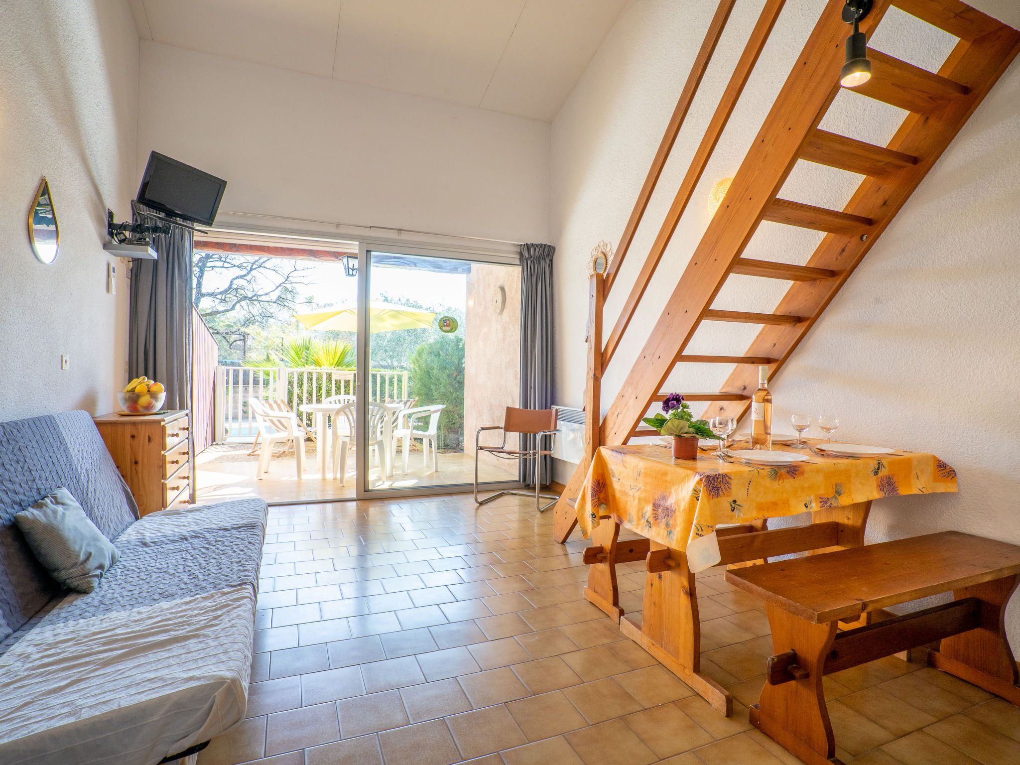 Ein gemütliches Zimmer mit einer Couch, einem Tisch mit gelber Tischdecke, einer Holzbank und einem Fernseher auf einem Schrank.