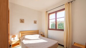 A bedroom with a wooden bed, a nightstand, a window with curtains, and a view of greenery outside. - Villa Marenco by Interhome (La Croix-Valmer)