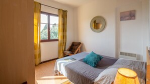 Een slaapkamer met een bed, een stoel, een raam met gordijnen, een ronde spiegel en een schilderij aan de muur.