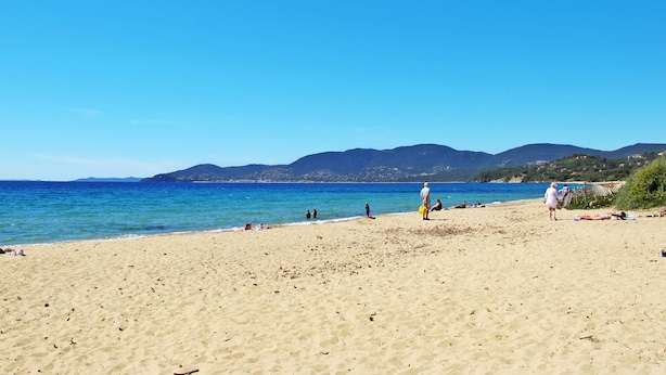 Een strand met helderblauw water, een zandstrand en bergen in de verte.