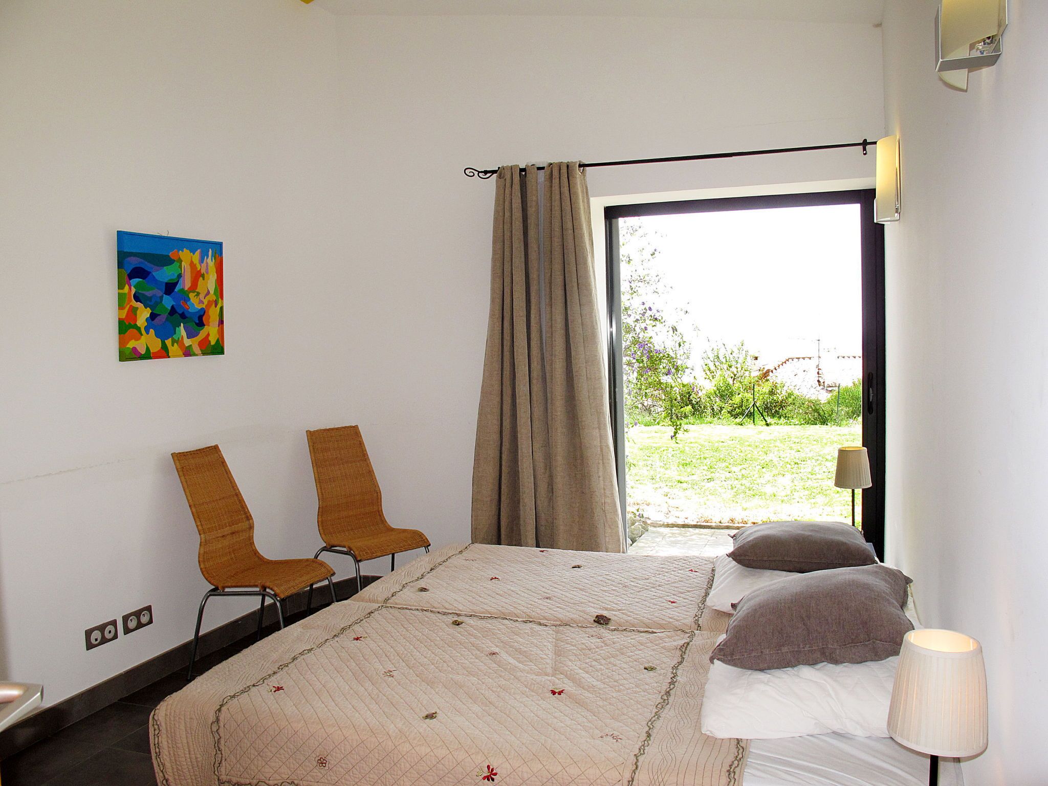 A hotel room with a bed, two chairs, a window with curtains, a painting on the wall, and a lamp.