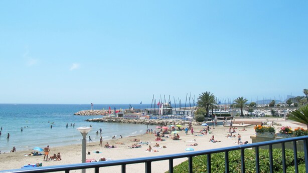 Une plage où des gens se prélassent au soleil et se baignent, une jetée ornée de drapeaux et des bateaux en arrière-plan.