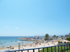 A beach with people sunbathing and swimming, a pier with flags, and boats in the background.