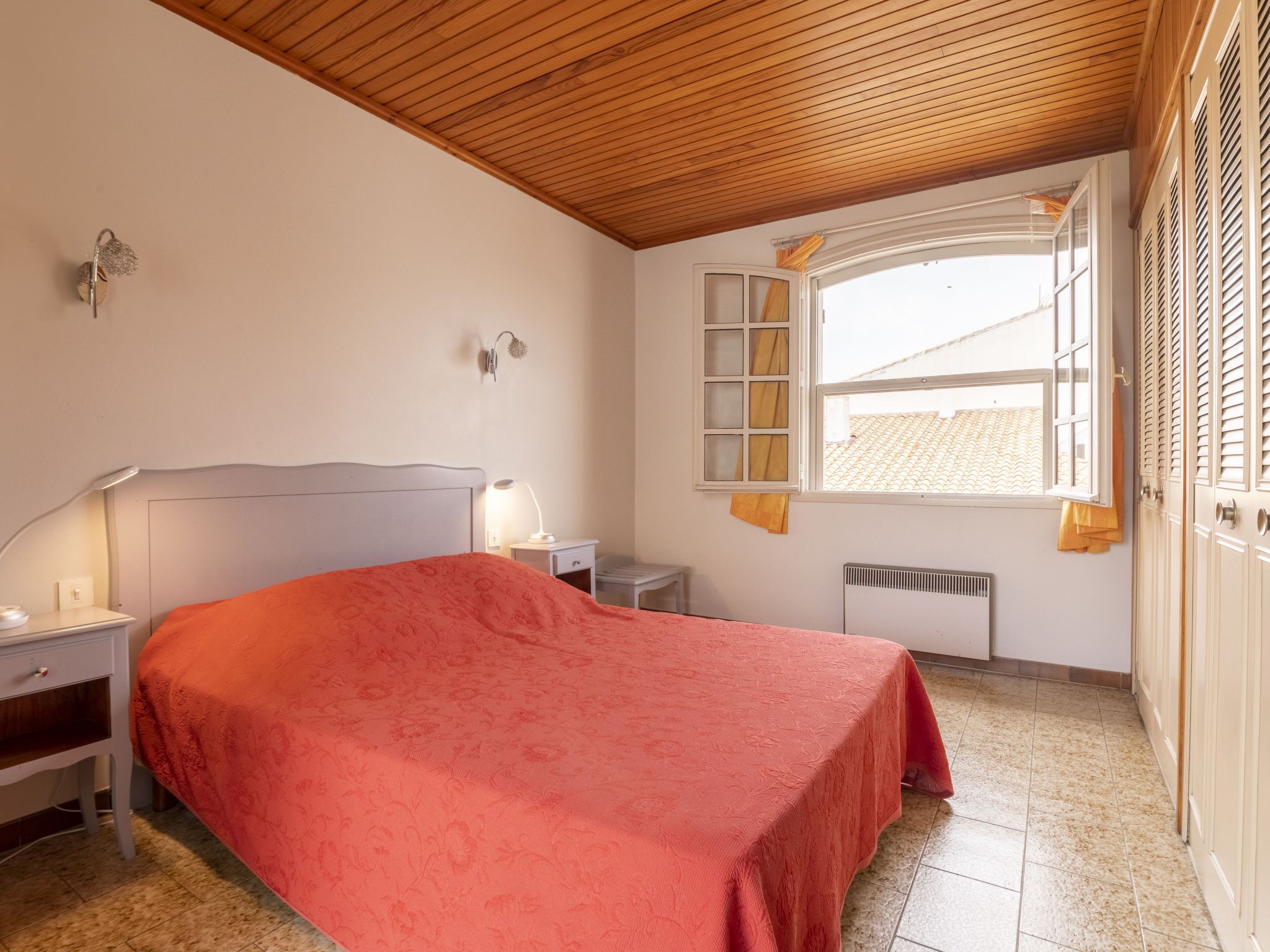 Una camera da letto con soffitto in legno, un letto con copriletto rosso, un comodino con lampada, una finestra con tende e un termosifone.