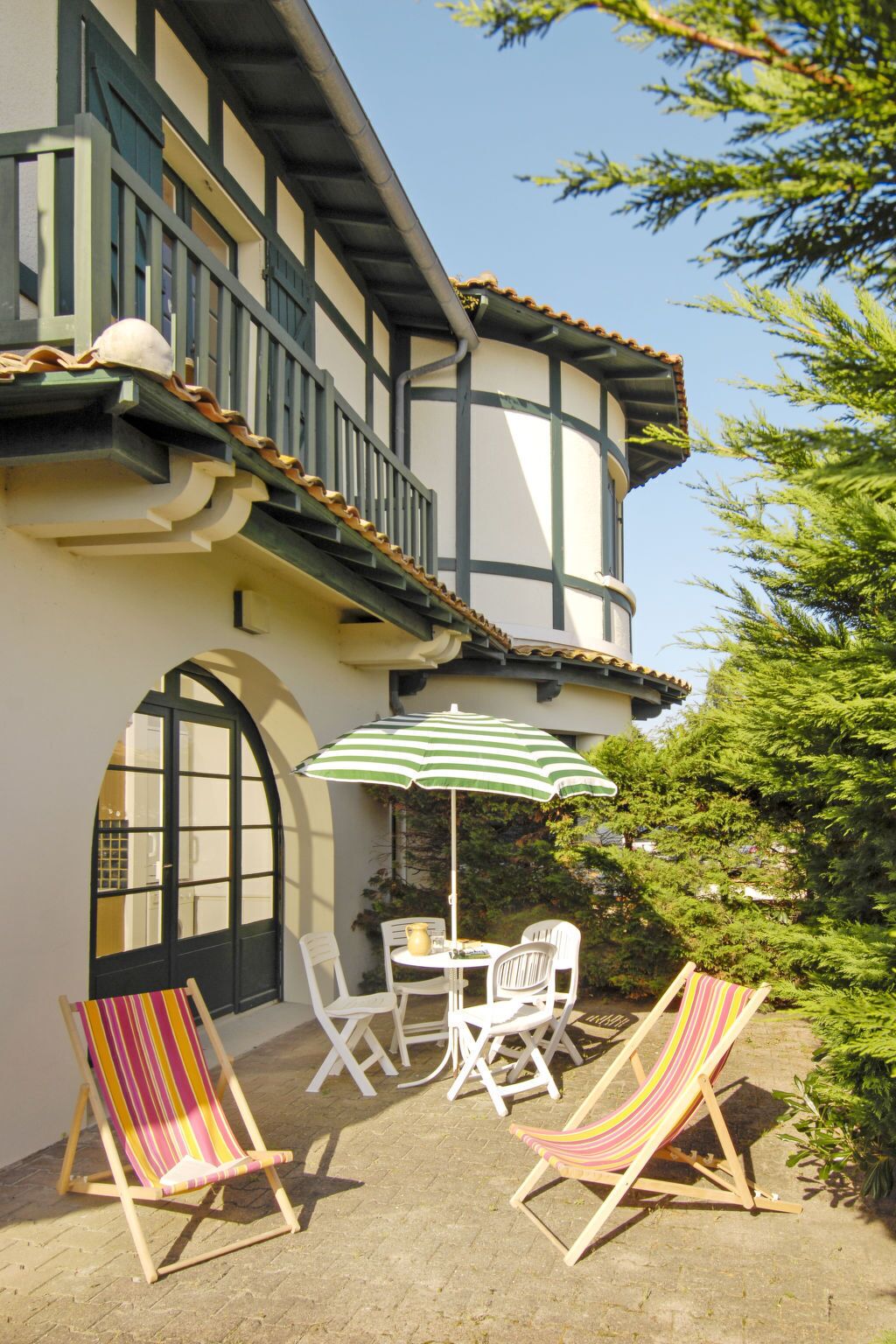 Un bâtiment à deux étages doté d’un balcon, d’une table à manger avec des chaises, et de deux chaises de plage à rayures.
