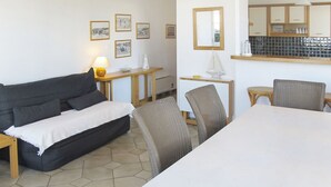 Une pièce comprenant un canapé, une table à manger, des chaises, une lampe, une petite table avec une lampe, ainsi qu’un coin cuisine avec des placards et une fenêtre.