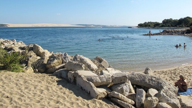 Une plage avec de gros rochers au premier plan, une étendue de sable et des personnes qui se baignent dans l’eau.