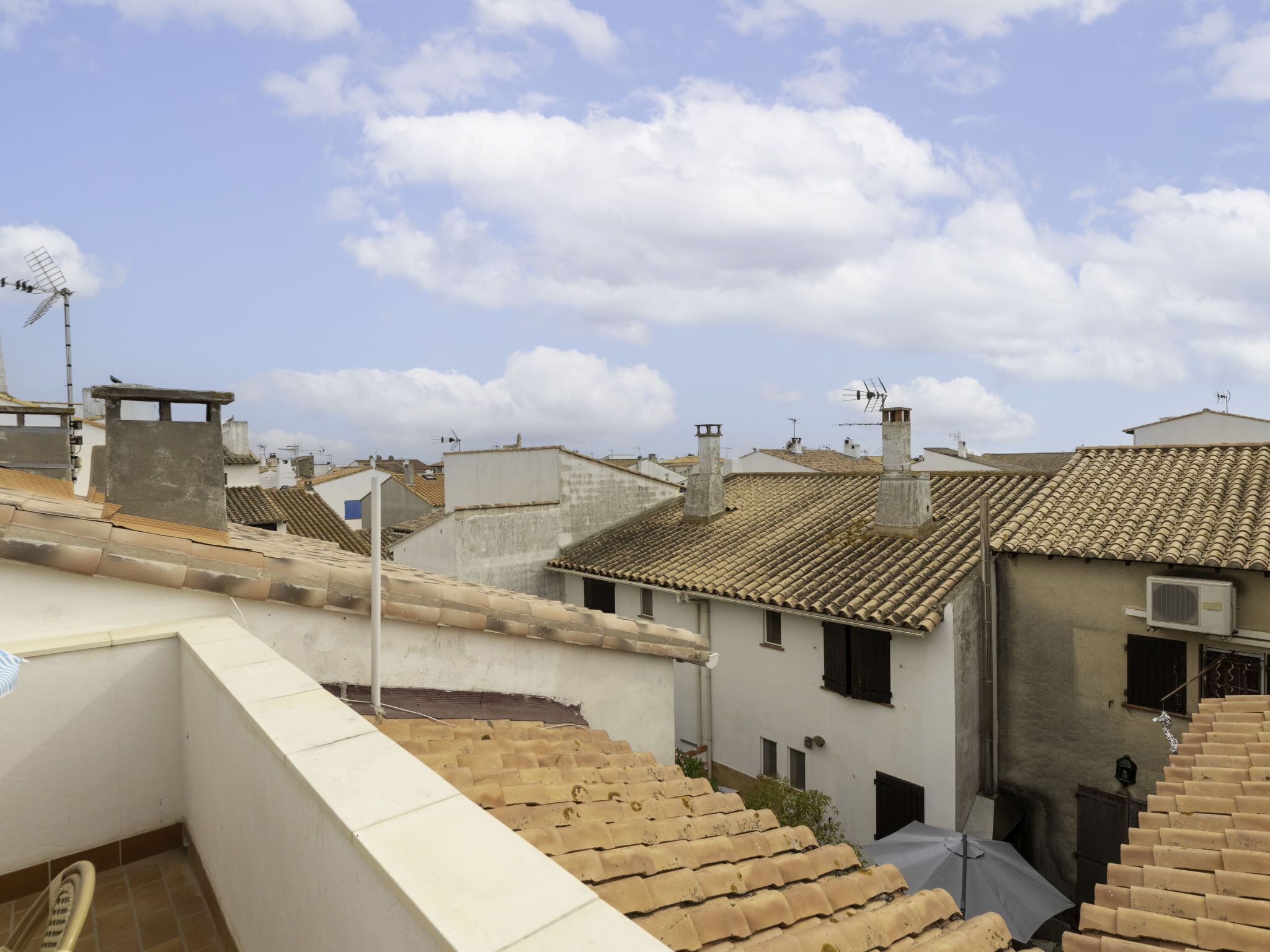 A rooftop terrace with terracotta tiles, a white building with a flat roof, and a clear blue sky with clouds.