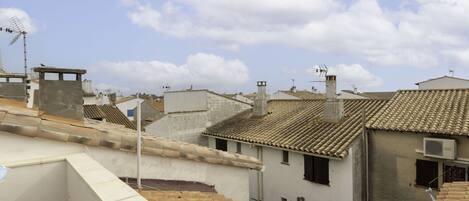 A rooftop terrace with terracotta tiles, a white building with a flat roof, and a clear blue sky with clouds.
