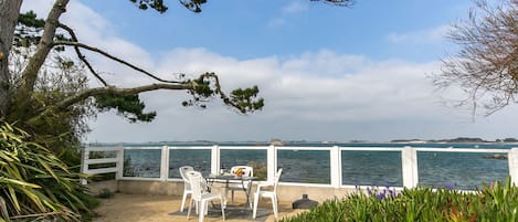 An outdoor seating area with a table and chairs overlooking a body of water, surrounded by greenery and a white fence.