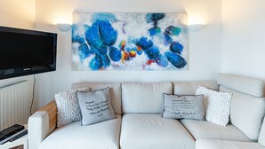 Ein Wohnzimmer mit einem weißen Sofa, zwei Zierkissen, einem Wand-Fernseher und einem farbenfrohen abstrakten Gemälde an der Wand.