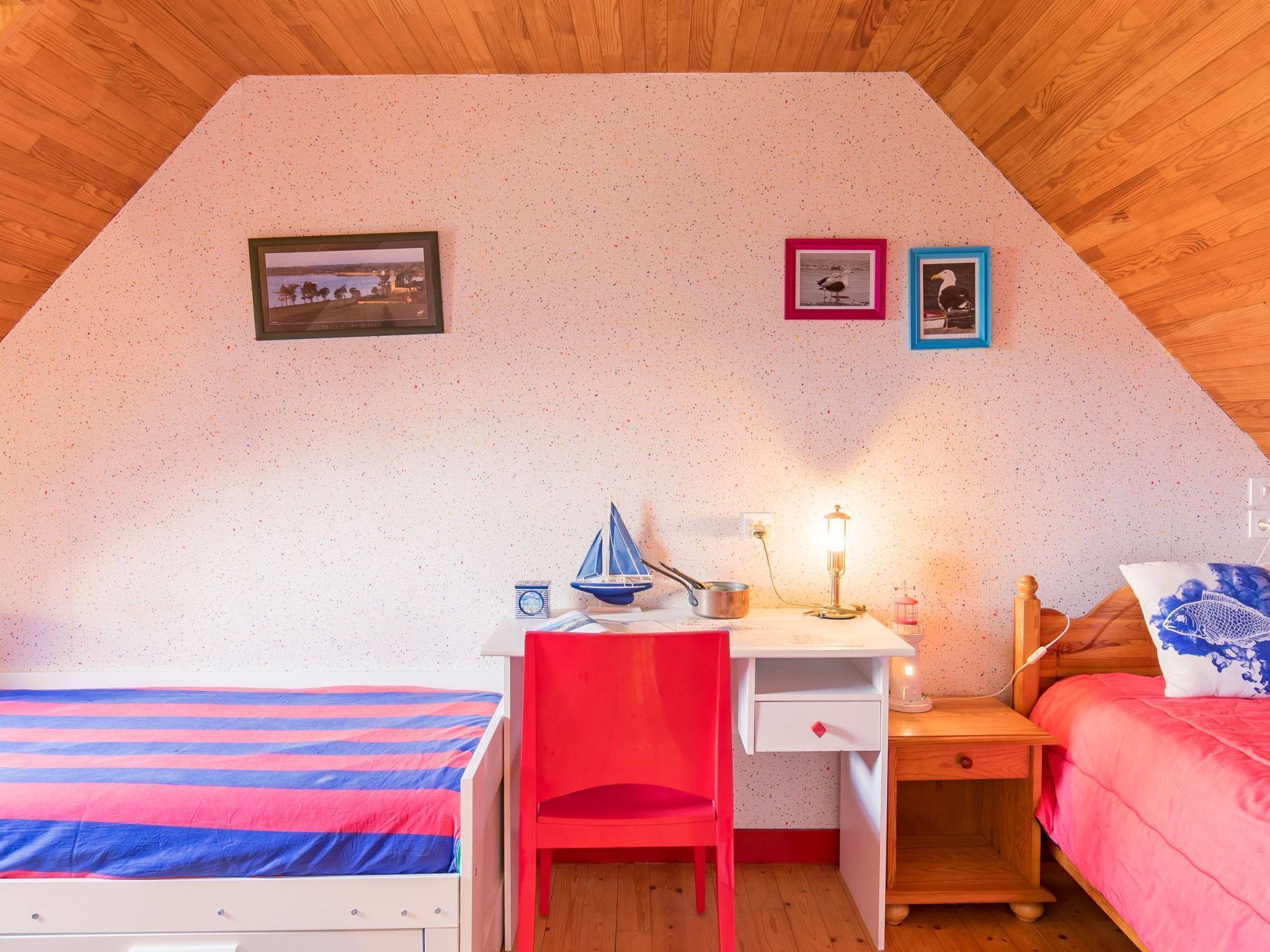 A bedroom with a sloped ceiling, a bed with a striped bedspread, a desk with a chair, a small table with a lamp, and two framed pictures on the wall.