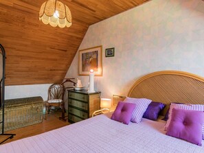 Een slaapkamer met een houten plafond, een bed met paarse kussens, een rieten stoel, een dressoir en een hanglamp.