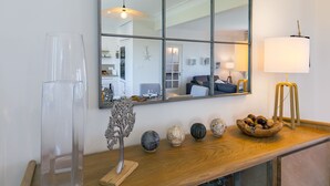 A modern living room with a large mirror, a wooden side table with decorative items, a standing lamp, and a glass cabinet.