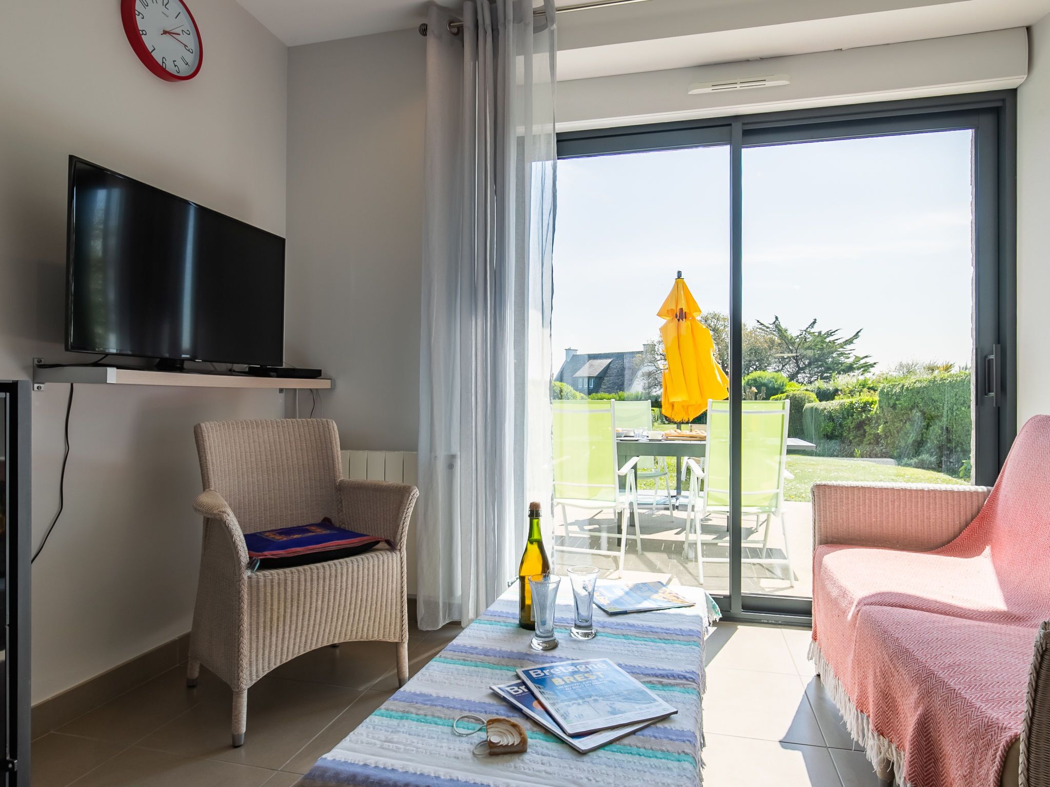 Ein modernes Wohnzimmer mit einem Flachbildfernseher, Korbstühlen, einem Tisch mit einer Zeitschrift und einer großen Schiebeglasdoor zum Balkon mit einem gelben Sonnenschirm.