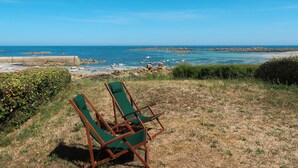 Dos sillones verdes en un área cubierta de césped con vista a una costa rocosa con barcos y un cielo azul despejado.