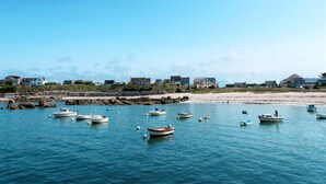 Escena costera con varias embarcaciones ancladas en aguas tranquilas, una playa arenosa y una hilera de casas al fondo.