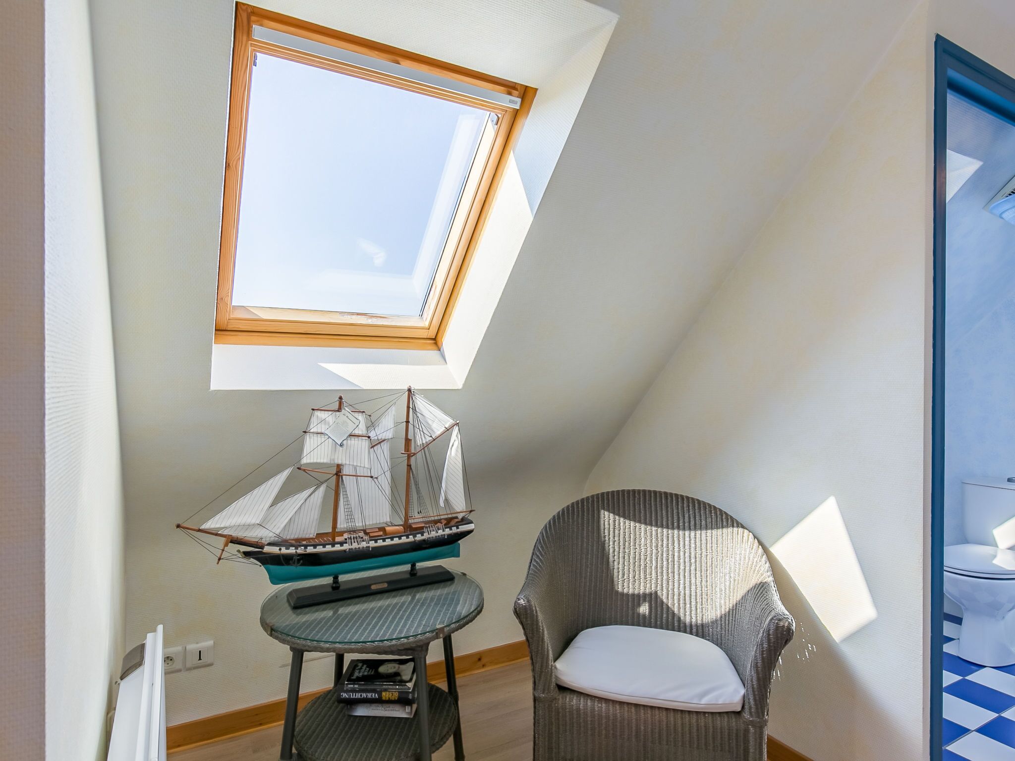Een kamer met een schuine kap, dakraam, model schip, rieten stoel en een kleine tafel.