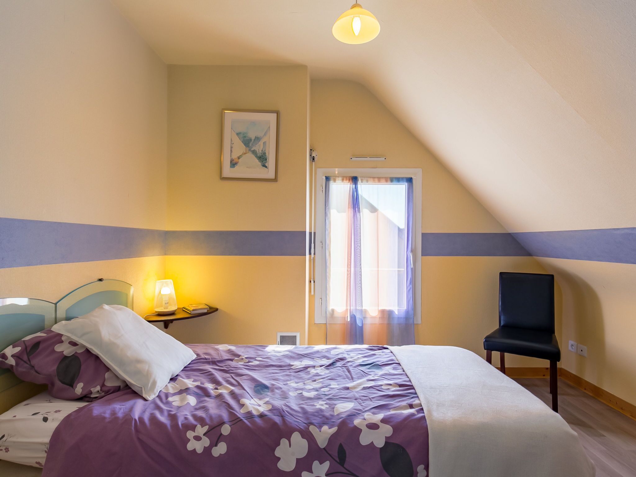 A bedroom with a bed, a chair, a bedside table with a lamp, and a window with curtains.