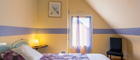A bedroom with a bed, a chair, a bedside table with a lamp, and a window with curtains.