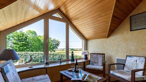 A cozy room with a sloped ceiling, wooden furniture, and large windows offering a view of greenery and a distant landscape.