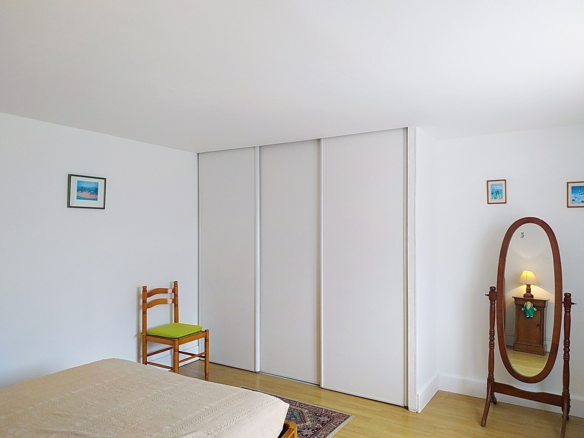 Ein Schlafzimmer mit einem Bett, einem Sessel, einem Kleiderschrank, einem Spiegel und zwei gerahmten Bildern an der Wand.