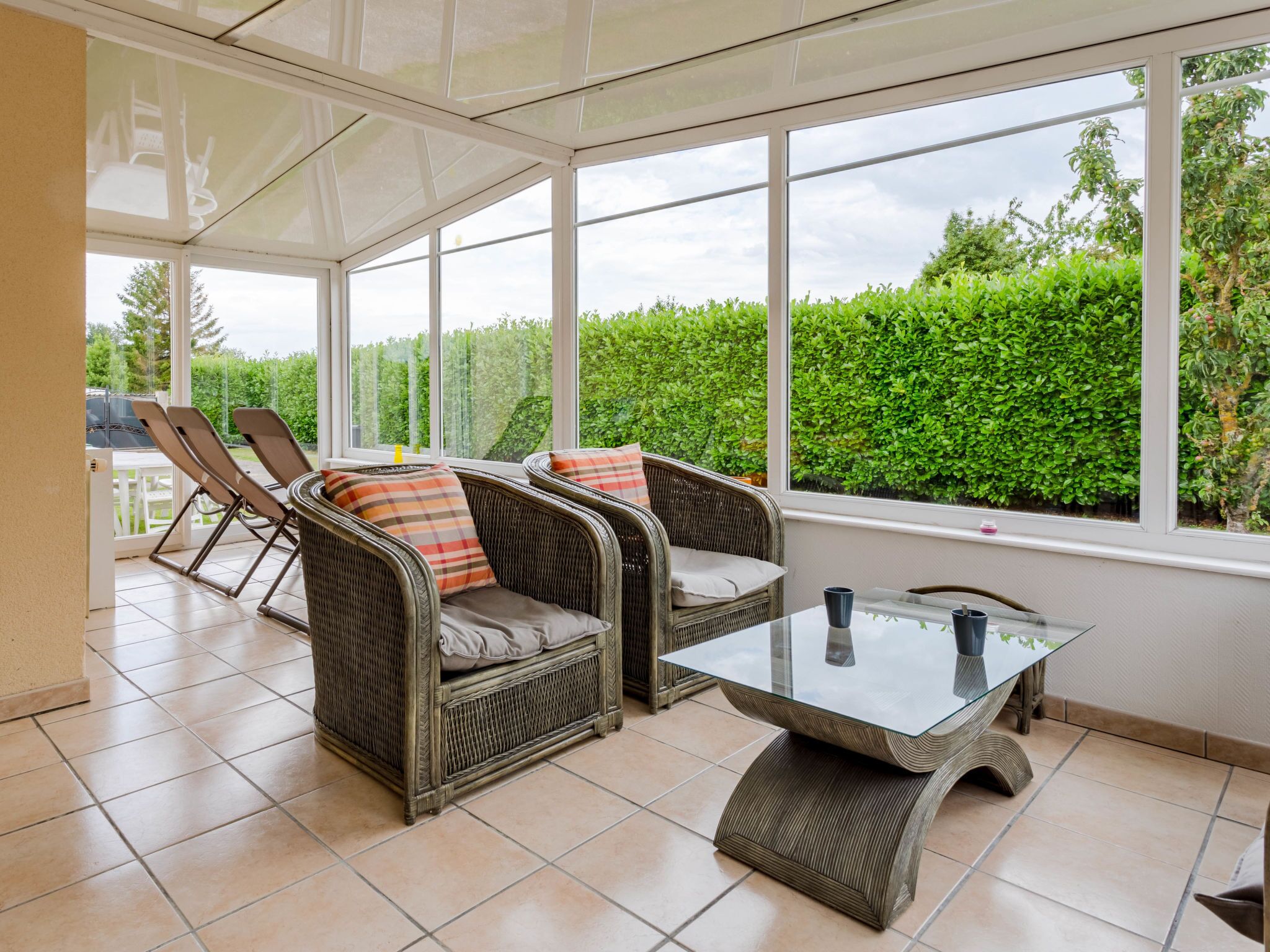 A sunroom with wicker furniture, a glass table, and a view of a green hedge.