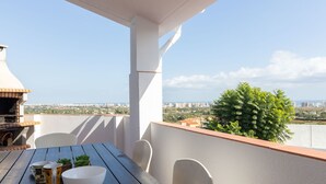 A balcony with a table, chairs, a grill, and a view of the city and sea.