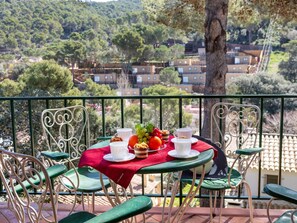 A terrace with a table set for two, featuring a red tablecloth, white cups, a plate of fruit, and a muffin. The terrace overlooks a village with buildings and trees. - S'Olivera Nº4 by Interhome (Sa Riera)