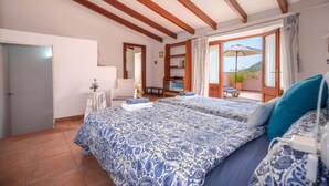 A bedroom with two beds, wooden beams on the ceiling, a small table with a vase, a chair, and a bookshelf. - Maria Fumet by Interhome (Capdepera)