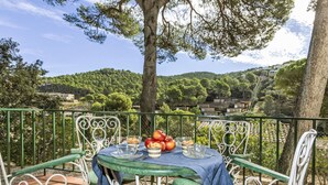A terrace with a table set for two, surrounded by green metal chairs, overlooking a lush landscape with trees and buildings. - S'Olivera Nº1 by Interhome (Sa Riera)