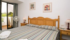Ein Hotelzimmer mit einem Bett, einem Nachttisch mit Lampe, einem Ventilator und zwei gerahmten Kunstwerken an der Wand.