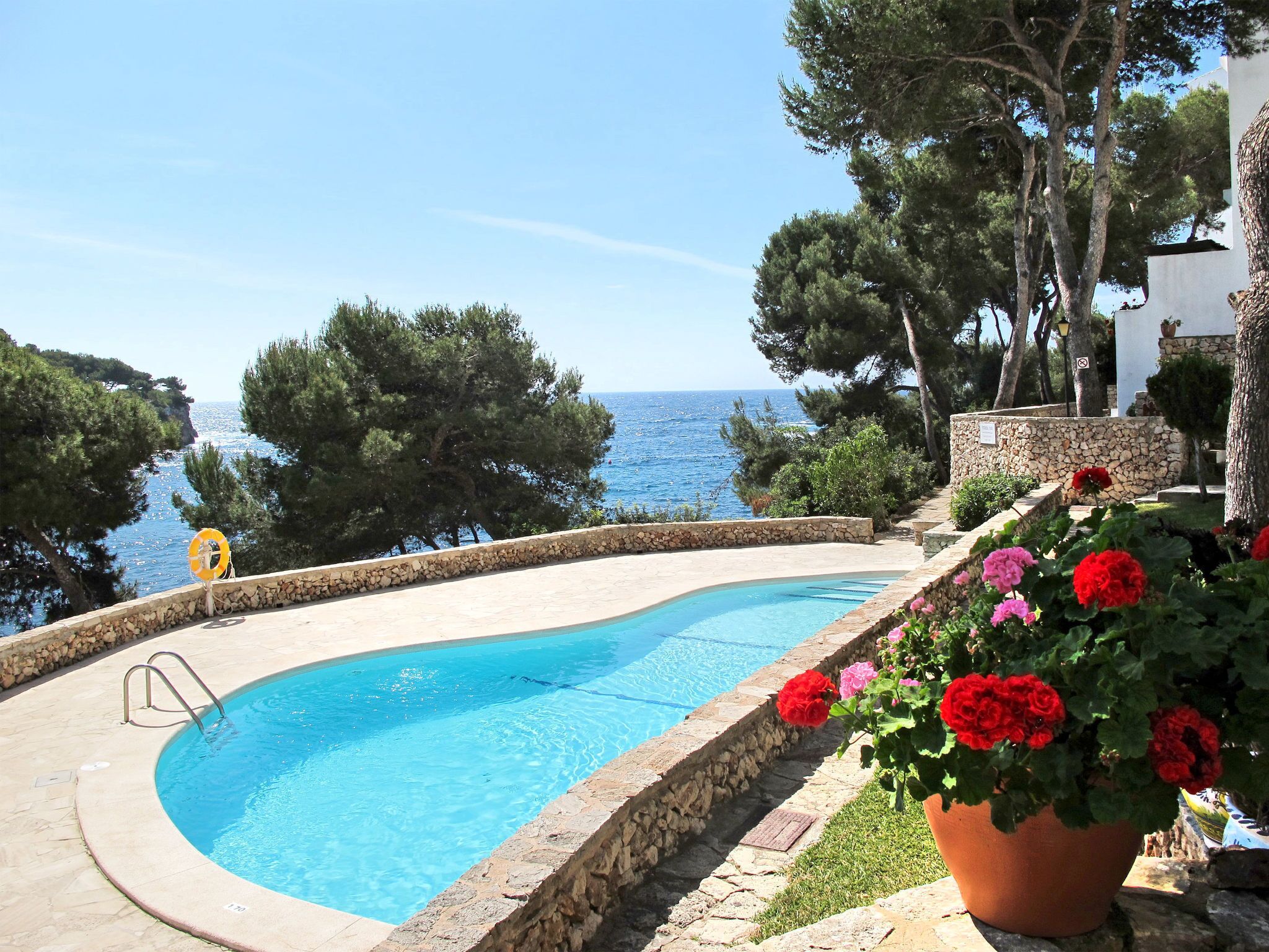 Ein geschwungener Pool mit Steinrand, umgeben von Kübelblumen und Bäumen, mit Blick auf ein Gewässer.