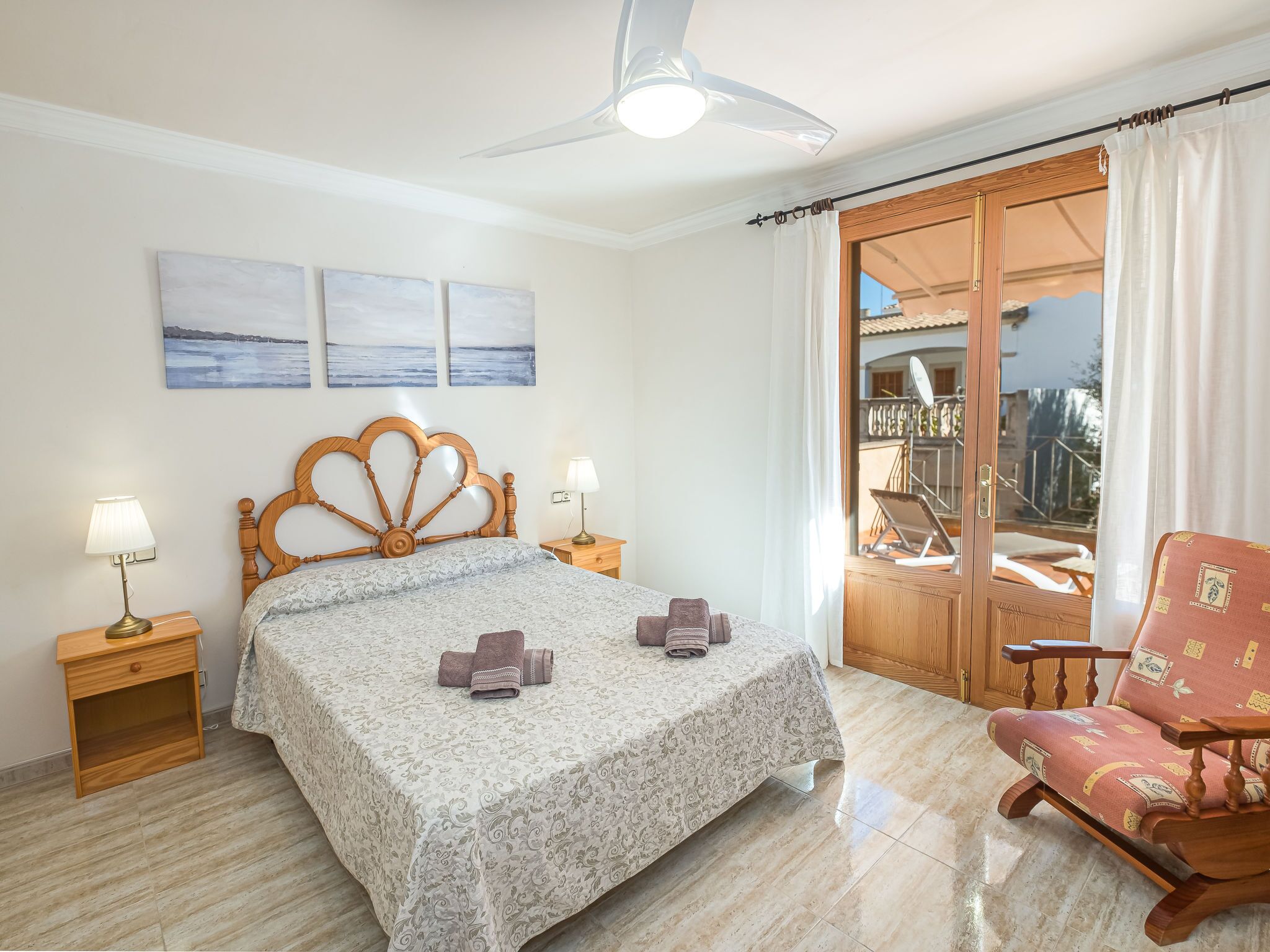Un dormitorio con una cama, dos lámparas de noche, una silla de madera, un ventilador de techo y vistas al balcón exterior.