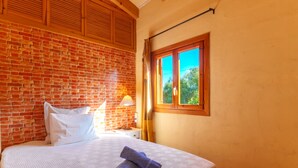 Una cama bien hecha con una colcha blanca y toallas enrolladas, un cabecero de madera, una pared de ladrillo, una ventana con vistas a la vegetación y un techo de madera.