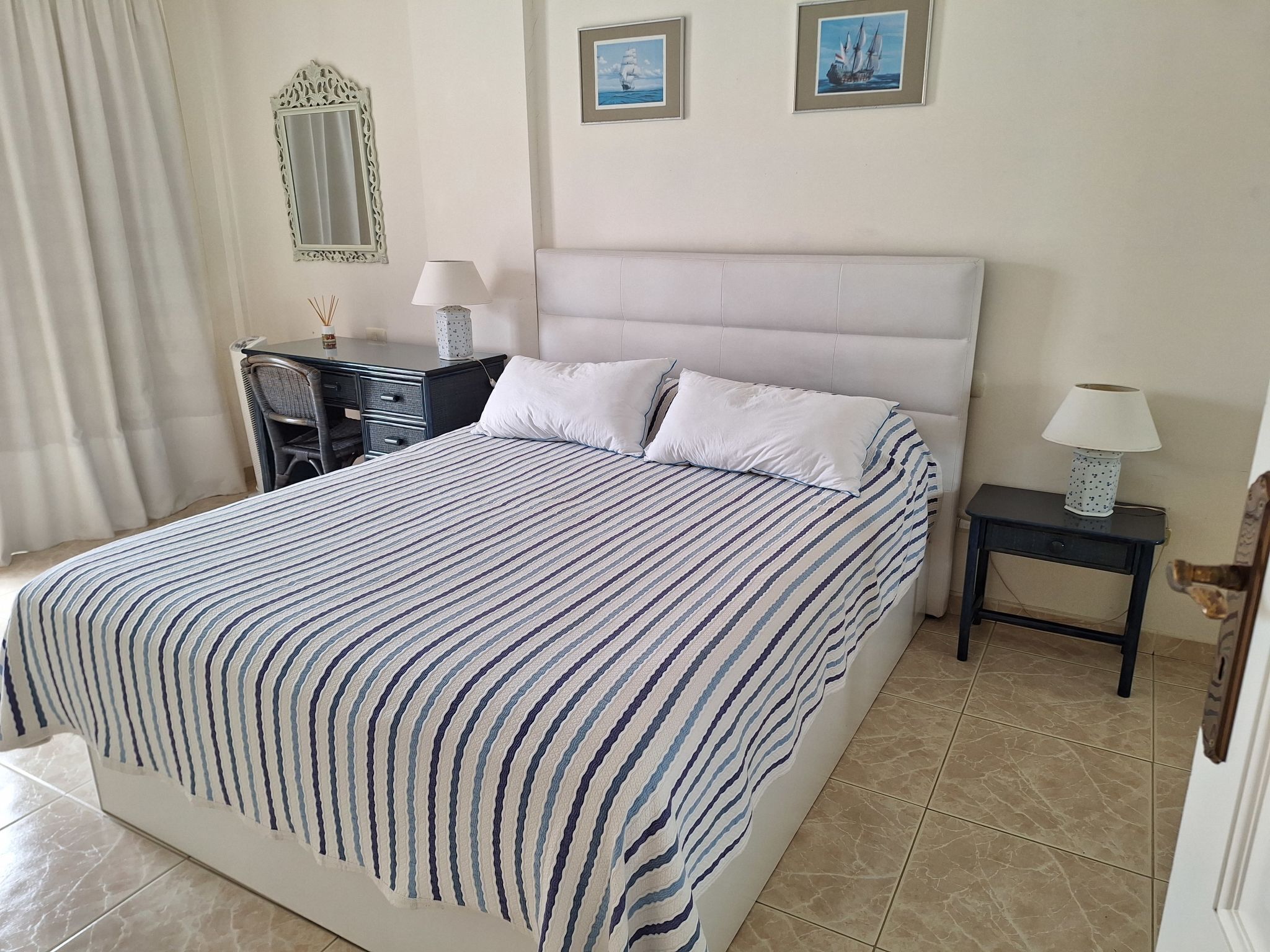 A neatly made bed with a striped bedspread, a bedside table with a lamp, a desk with a chair, and a mirror on the wall.