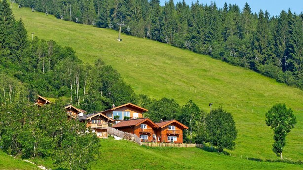 Un chalé de madera con balcón, rodeado por una ladera verde exuberante y un telesilla al fondo.
