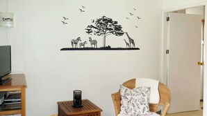 A living room with a television, a wicker chair with patterned cushions, a small wooden side table, and a wall decal of a tree and animals.