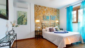 Un dormitorio con pared de piedra, una cama con ropa de cama blanca, una mesita de noche, una lámpara de pared y una ventana con cortinas azules.