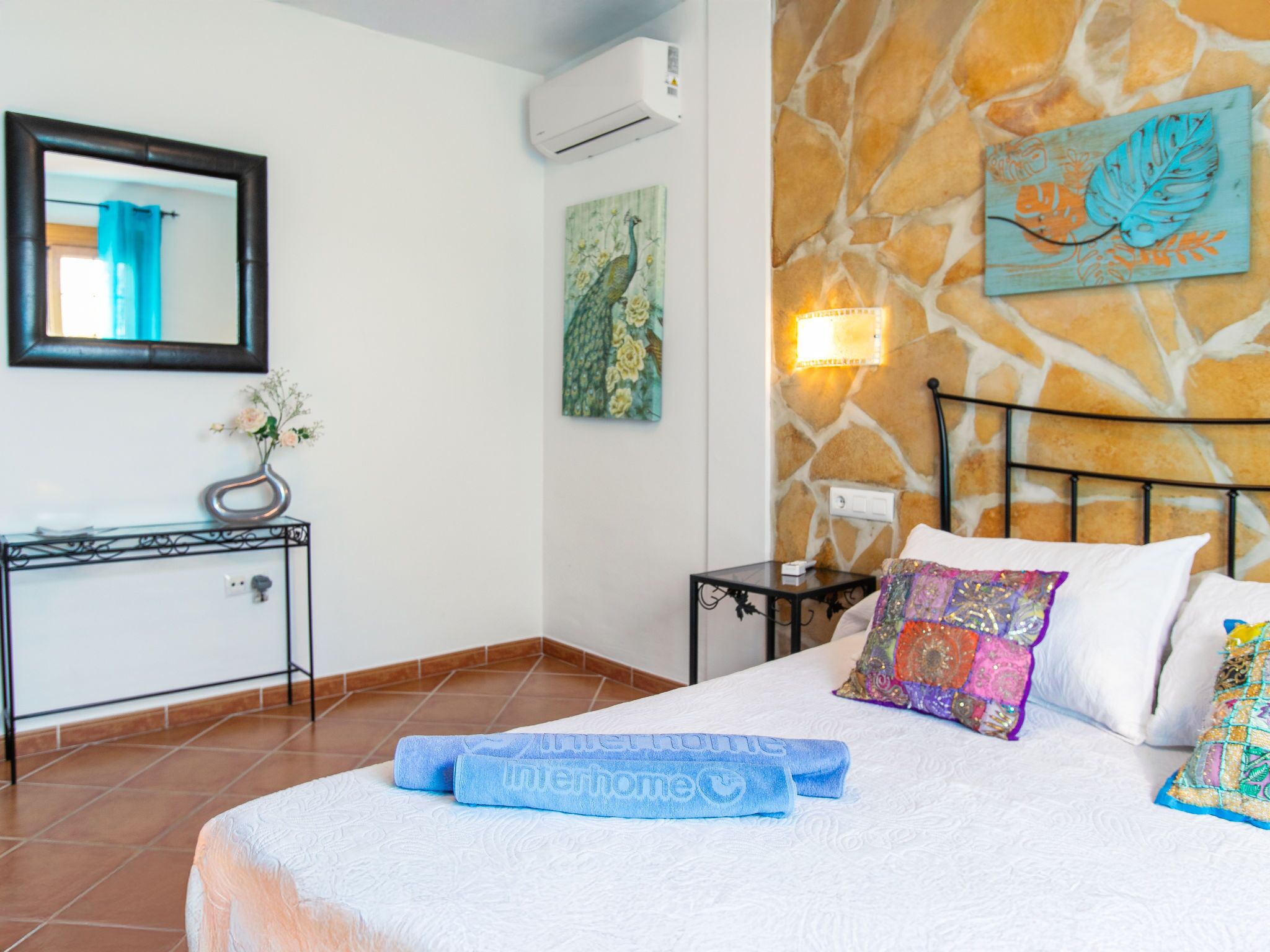 A hotel room with a bed, a nightstand, a mirror, a wall-mounted air conditioner, a painting, and a decorative wall piece.