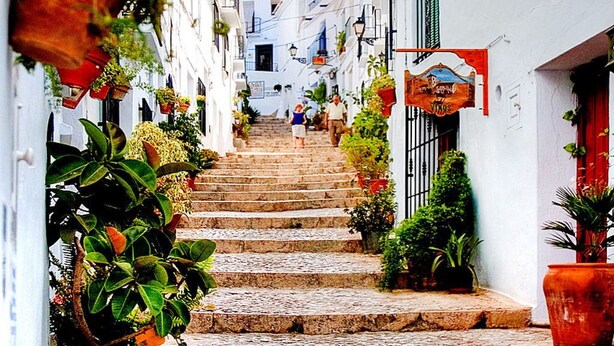 Una calle empedrada con edificios blancos, plantas en macetas y un cartel de un negocio local.