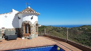 Zona de piscina con vista al mar, un edificio blanco con detalles de piedra y mobiliario exterior.