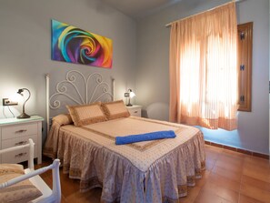 Quarto com cama, duas mesinhas de cabeceira, um criado-mudo, cabeceira decorativa, pintura abstrata colorida e janela com cortinas.