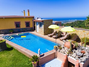 Piscina com espreguiçadeiras e guarda-sóis, cercada por um muro de pedra e plantas em vasos.