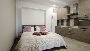 Un dormitorio moderno con cama, mesita de noche, lámpara y una kitchenette con armarios y electrodomésticos.