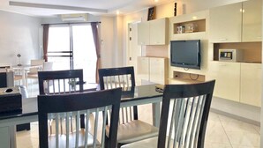 A modern living room with a dining area, featuring a flat-screen TV, built-in cabinets, and a large window with curtains. - 1 Bedroom Seaview 706 Kiengtalay (Pattaya)