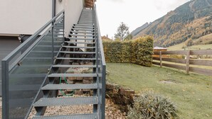 A modern staircase leading to a building with a metal railing, a grassy area with a wooden fence, and a mountainous backdrop.