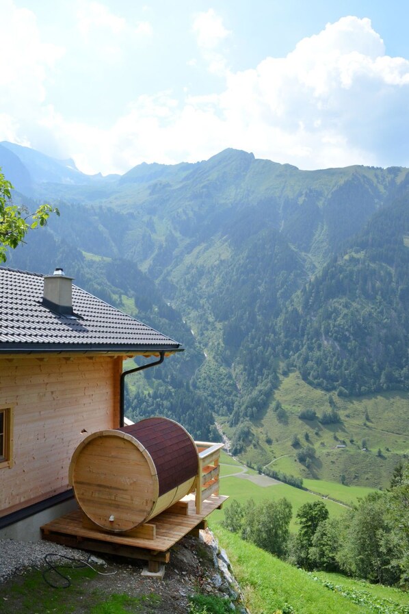 A wooden cabin with a large barrel on a wooden platform, overlooking a valley with mountains in the background.