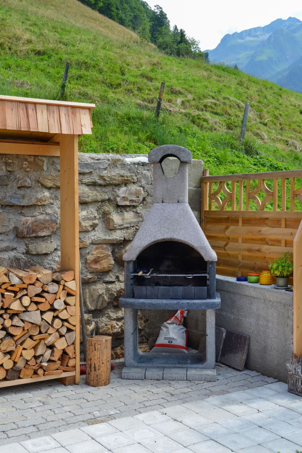 An outdoor stone pizza oven with a wooden roof, a stack of firewood, and a stone wall.